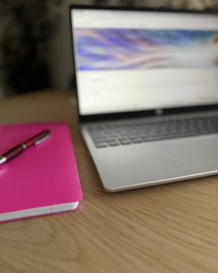 Pinkes Notizbuch mit Stift auf einem Holztisch, daneben ein Laptop